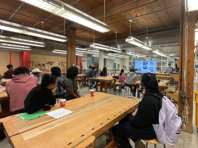 Multiple students seated at wooden desk in a workshop style classroom looking towards the front and listening to a presentation. 